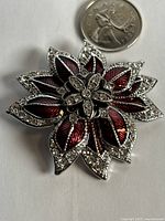 Close-up of Monet floral brooch with silver-tone edges, red enamel leaf petals and clear rhinestones, with Canadian quarter coin next to it for size reference.