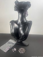 Black display bust showing one pair of purple and iridescent dangle earrings worn; the other earring pair and brooch shown at base along with a small bag of extra beads.
