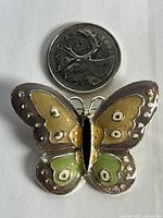 Front view of vintage butterfly brooch next to a Canadian 25 cent coin for size reference. The brooch is enamel with gold tone and tan, green, black colors.