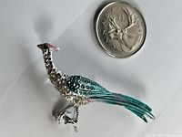 Photo showing the vintage roadrunner bird brooch pin next to a Canadian 25 cent coin for scale, highlighting the silver tone metal, rhinestone embellishment, and turquoise and pink enamel details.