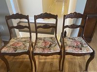 Four wooden dining chairs lined up side by side showing the floral cushioned seats and carved backs.