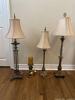 Four metal lamps standing side by side on hardwood floor with beige walls and white baseboard in background.