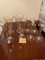 Photo shows four large amber glass goblets with clear stems and bases and four smaller amber glass candlestick holders with clear bases arranged on a dark wooden surface.