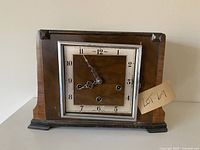 Front view of antique wooden mantel clock showing metal face with Arabic numerals and decorative clock hands. Clock face has three winding holes, door missing glass.