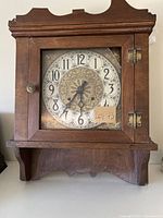 Front view of antique wooden mantel clock with a decorative circular dial behind a glass door. The wood shows signs of fragility and the top right corner of the glass is broken.