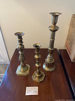 Three brass candle holders of varying heights on a wooden surface, showing hexagonal and rounded bases and detailed shafts.