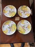 Four round dessert plates with a colorful design of lemons, blossoms, and butterflies on a white background, arranged in a square formation on a wooden surface.