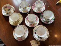 Overview of all 8 china teacup and saucer sets arranged on a wood table showing floral patterns and gold accents