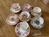 Photo of 7 fine china teacups with saucers showing floral and gold decorative patterns, arranged on wooden table.