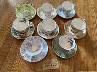 Photo showing all 8 china teacups with matching saucers arranged on a wooden surface, each with distinct floral and gold trimmed designs.