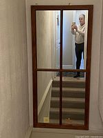 Full frontal photo of a large rectangular country style mirror with a wooden frame divided into four sections by wooden muntins. Mirror is mounted on a wall above a staircase.
