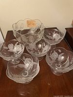 Five clear glass dessert dishes with etched poppy flowers and one large glass bowl on wooden surface.