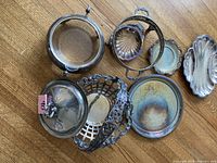 Top view of all silver items arranged on wooden floor showing magnifying glass stand, decorative basket, shell dish, tarnished round tray, and small spoons/dishes.