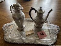 Silver set including two vessels (water jug and teapot) displayed on decorative rectangular platter wrapped in plastic.