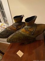Pair of tin decorative containers shaped like boots with worn metallic finish and chain detail around ankles, shown from front angle.