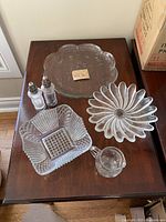 Six glassware items arranged on a wooden table: a round etched plate, fluted bundt pan, square divided dish, small creamer jug, and a pair of vintage salt and pepper shakers.