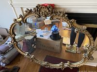 Full view of the large Victorian-style mirror showing the oval shape and highly ornate, gold-toned carved frame with floral and scroll decorations.