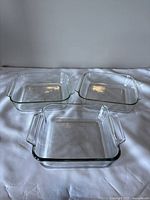 Three square glass baking dishes arranged on flat surface; two are identified as Pyrex, all clear transparent glass with handles.