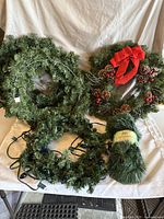Three artificial Christmas wreaths including one with pine cones and red bow, one plain wreath, one wreath with blue lights plus a coiled roll of artificial garland.