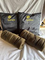 Front view of two black Bushtukah camping pillows showing embroidered yellow logo and two rolled olive green camping blankets in front.