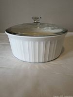 White circular Corningware baking dish with clear glass lid on top, displayed against plain background.