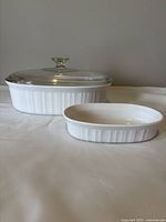 Set of two oval Corningware baking dishes, one with a glass lid (larger) and one without (smaller), shown side by side on a light surface.