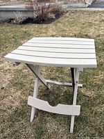 Beige folding side table unfolded standing on grass showing slatted top surface and foldable legs.