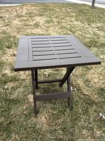 Front view of small brown folding plastic side table outdoors on grass, showing square slatted tabletop and folding legs.