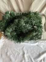 Green artificial pine needle garland coiled loosely in a plastic container, showing the texture of the needles.