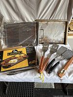 Photo of grilling utensils including spatulas, tongs, perforated grill plates, and GrillPro griddle box on white cloth background.