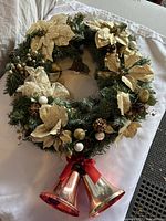 Overall view of the large green holiday wreath decorated with gold poinsettias, berries, pine cones, white balls, and two hanging bells tied with a red ribbon.