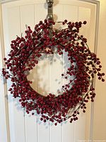 Front view of the red berry wreath hanging on a white door, showing dense clusters of red berries on brown branches arranged in a circular shape.