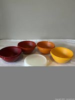 Set of four Tupperware plastic bowls in red, dark orange, bright orange, and yellow with a white lid in front, displayed on white surface.