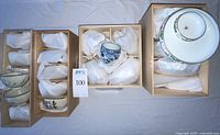 Four wooden boxes containing Asian porcelain cups and bowls, with one Koransha set unwrapped for photo. Each set is wrapped in cloth inside boxes.