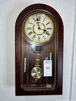 Front view of wooden mechanical wall clock, Roman numeral dial visible with two brass weights and pendulum.