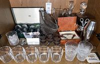 Full view of barware collection including decanters, rocks glasses, cut glass tumblers in clear and aubergine, wine glasses in box, wire basket, and bar tools on wooden shelf.