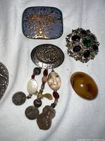 Top view of various brooches and pins including tree motif square brooch, oval brooch with dangling elements, ornate gemmed brooch, and amber cabochon pin.