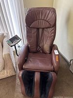 Front view of the brown massage chair showing padded seat, backrest, foot massage section, and attached remote control panel.