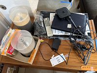 Photo showing pile of electronic devices including Toshiba DVD player, Apple TV, multiple cables, remote controls, and DVDs in empty spindle cases and original packaging.