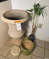 Large faux ceramic urn style planter beside green ceramic pot, bamboo plant holder, saucer, and plant elevators on tile floor