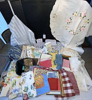 Wide view of assorted table linens including embroidered white tablecloth, floral patterned napkins, checked cloths, and other assorted kitchen textiles.