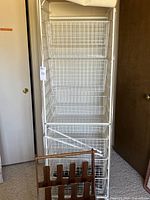 Full view of the tall white metal wire frame storage unit with six canvas-covered wire baskets on casters, standing upright against the wall, with a wooden shoe rack placed in front.