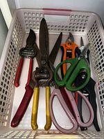 Photo of various pruning shears and scissors with plastic and metal handles in a white plastic basket