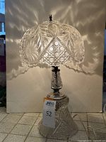 Full view of the lamp lit, showing the intricate etched glass shade casting patterned light and the clear glass base with metal accents and finial.
