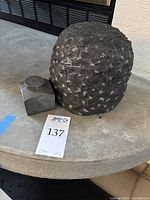 Photo showing the rough textured dark stone sculpture and the small square metal jar with lid side by side on a round concrete surface with lot number card.