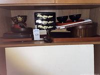 Shelf view with stacked lacquerware items including trays, bowls, boxes, and bamboo mats.