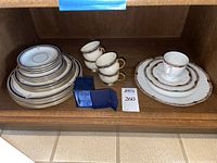 Display of stacked Lenox Buchanan plates and cups alongside Tirschenreuth Palais plates and cups with blue tape above shelf