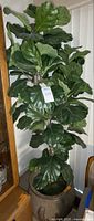 Artificial fiddle leaf fig tree approximately 6 feet tall with broad dark green leaves. Tree is inside a pot and the plant looks whole.