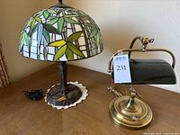 Pair of lamps on wooden surface. Left lamp is Tiffany style with green and yellow bamboo leaf stained glass shade and metal base. Right lamp is classic desk lamp with dark green glass shade and brass base.