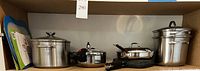 Wide shot of shelf containing large stainless steel pot with glass lid, double boiler, pressure cooker, and plastic cutting boards on left side.